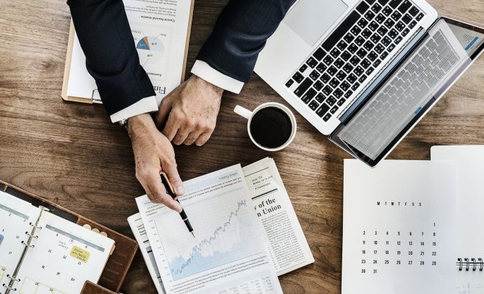 Photo of a table with reports, laptop, coffee and the arms of a man in a suit