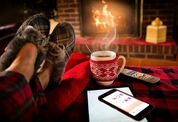 Feet in slippers resting on a table with a warm beverage, phone, remote control in front of a fireplace.