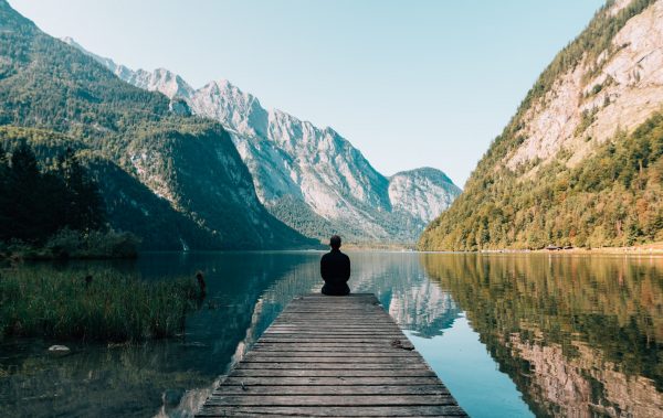 Una persona sentada al borde de un embarcadero en un lago rodeado de montañas