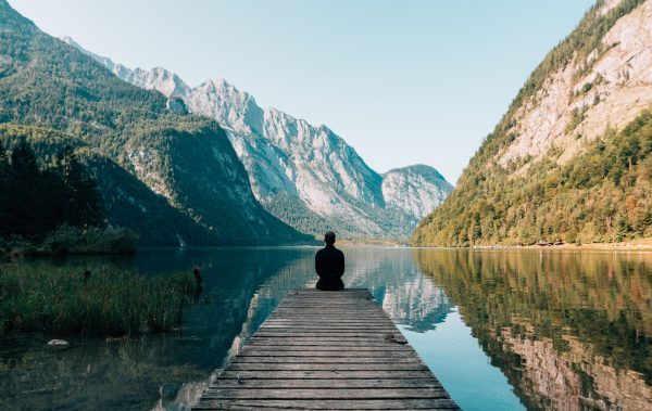 A person sitting at the edge of a dock on a lake surrounded by mountains